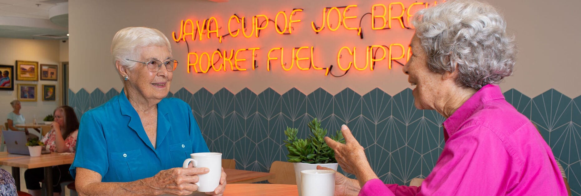 Two senior women laughing and talking over coffee at a table inside Posada Java.